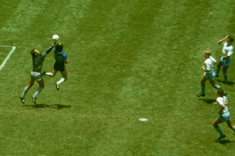 A soccer star in a blue and black kit outjumps a goalkeeper with his arm outstretched. England players in white look on.
