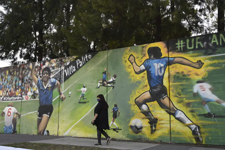 A woman wearing black walks past a wall with images of Diego Maradona running.