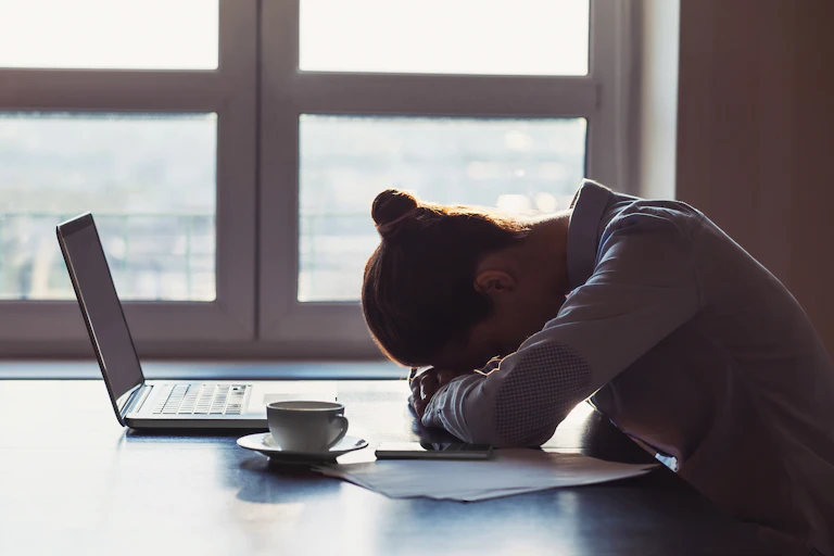 A woman sitting at a desk with her head resting on her folded arms