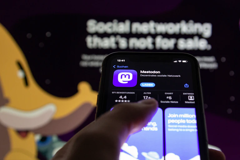 a phone screen showing the Mastodon with a screen in the background with the text SOCIAL NETWORKING THAT'S NOT FOR SALE