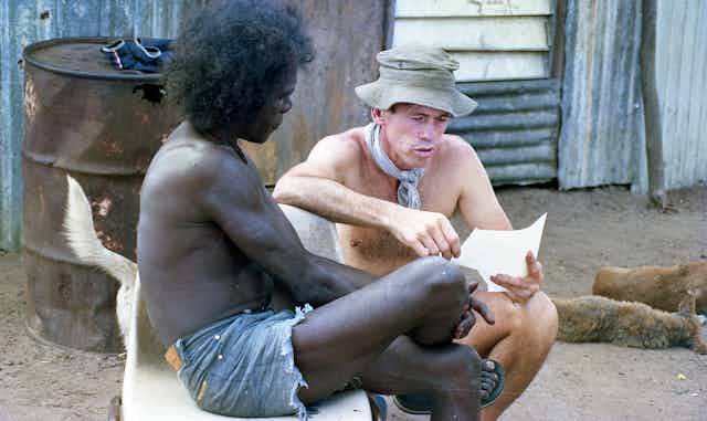 A Yolngu man sits on a bench, talking to a white man in an army hat, both shirtless in shorts. A dog is in the background