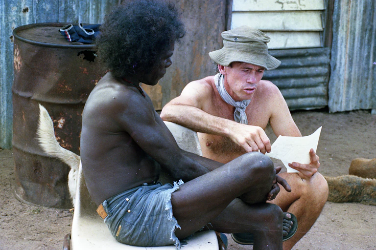 A Yolngu man sits on a bench, talking to a white man in an army hat, both shirtless in shorts. A dog is in the background