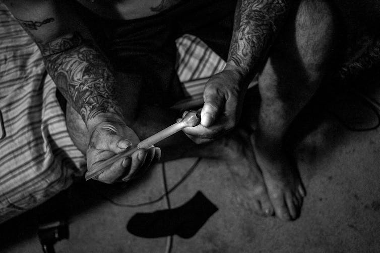 Black and white overhead view of a man's hands lighting a broken glass meth pipe. His arms are tattooed, and his bare feet are visible on the floor.
