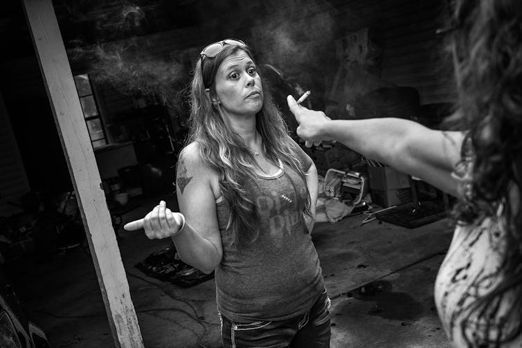 Michelle, a woman with long wavy hair wearing a tank top, stands in front of an open garage. Jennifer, who is mostly out of frame, holds out a cigarette towards Michelle.