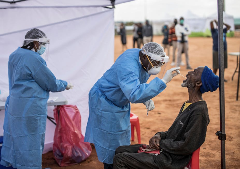 Two medical professionals in scrubs take a throat swab of a man under a tent in a dry landscape in Africa. A line of people wearing face masks waits to be next for testing.  
