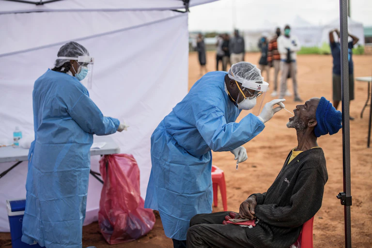Two medical professionals in scrubs take a throat swab of a man under a tent in a dry landscape in Africa. A line of people wearing face masks waits to be next for testing.