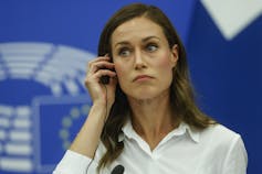 A woman with long dark hair and wearing a white blouse adjusts her earphone.