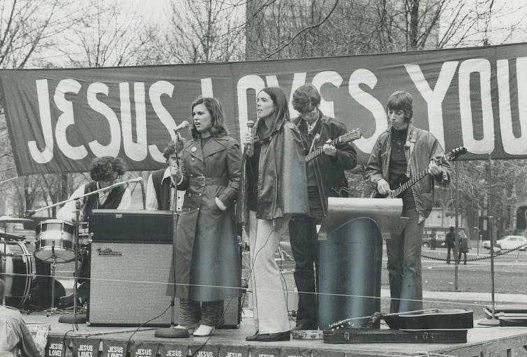 A black and white photo shows a small group of musicians on a stage in front of a banner saying 'Jesus loves you.'