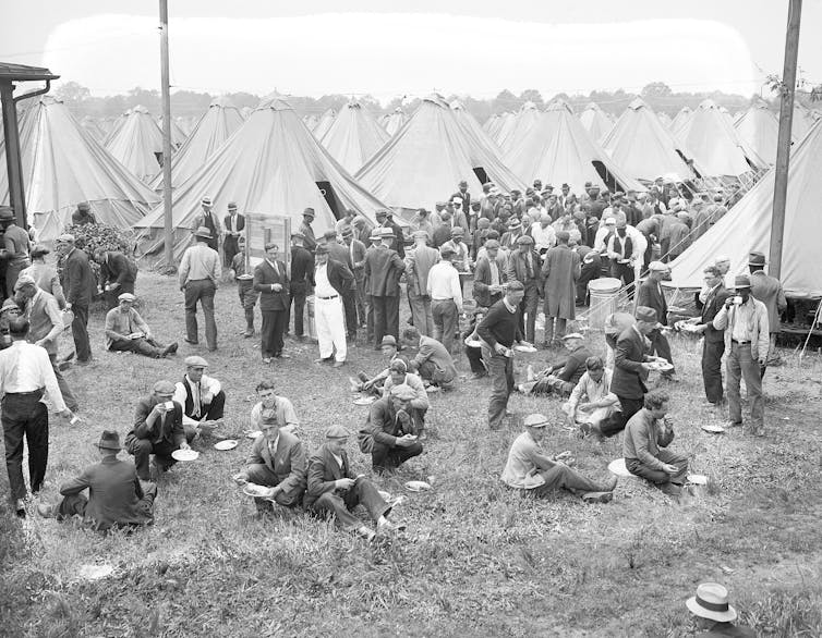 Groups of men eat lunch while sitting and standing near dozens of tents.