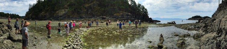 People stand near a shoreline in front of a curved line of rocks. A woman stands behind the rocks speaking to the people.