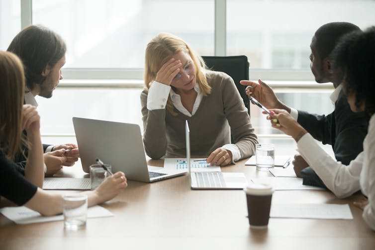 A woman sits at the head of a group of people with her head in her hand. The people surrounding her are aggressively gesturing toward her