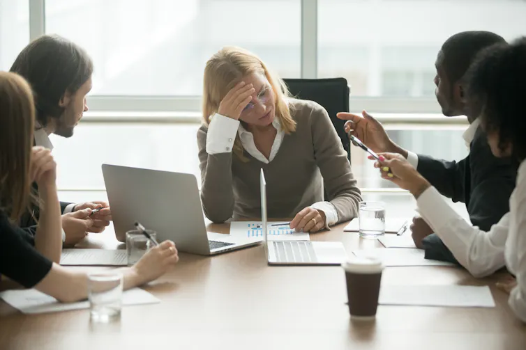 A woman sits at the head of a group of people with her head in her hand. The people surrounding her are aggressively gesturing toward her
