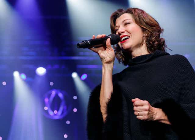 A brunette woman with red lipstick wearing a black poncho sings into a microphone. 