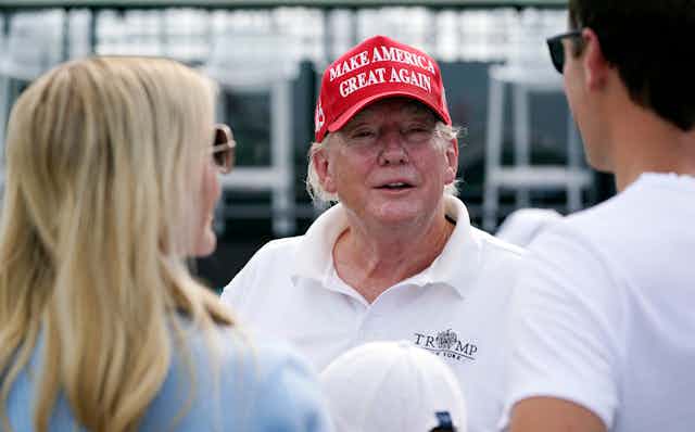 An aging man in a red hat that says make america great gain and a white t-shirt talks to two people.