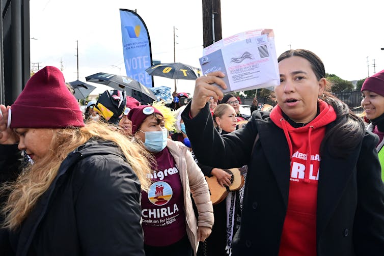 A brown skinned young woman holds up a ballot and stands amid a crowd of people outside.