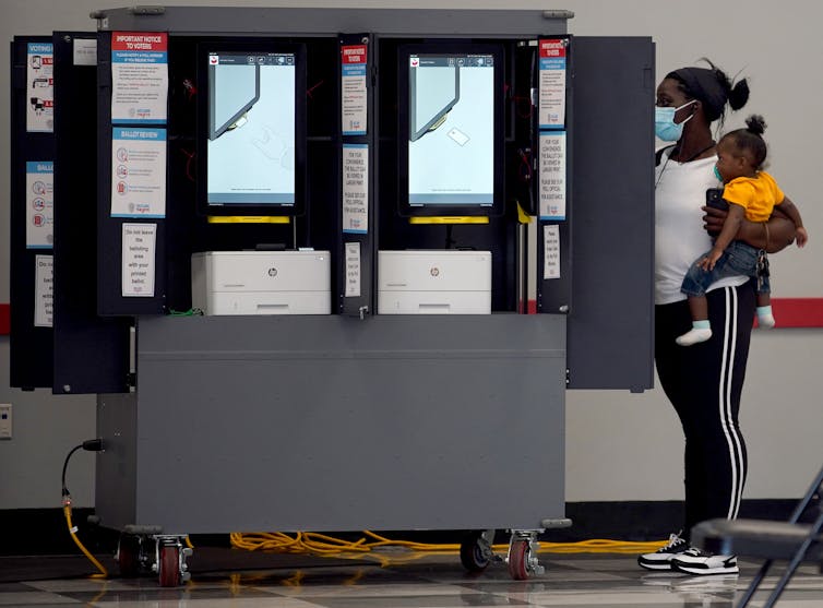 A young Black woman holds a baby on her hip and votes at a shielded voting booth.