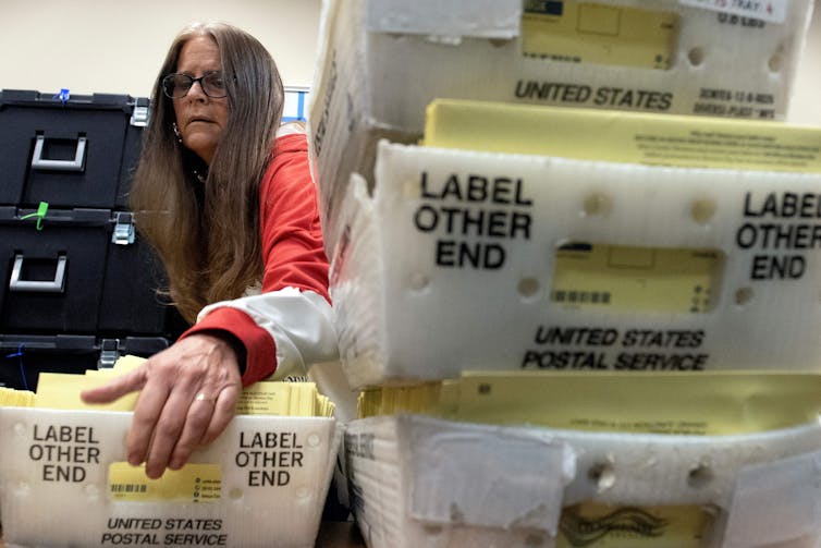 A middle aged white woman with long brown hair appears to pick up a white box that has yellow envelopes inside. Next to her sits a pile of more white boxes with yellow envelopes.
