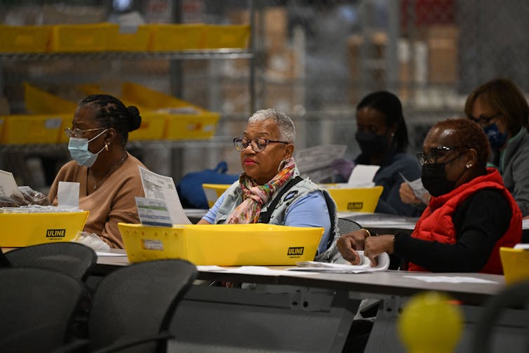 Two rows of five Black women sit at long tables, sorting through papers that are in yellow boxes.