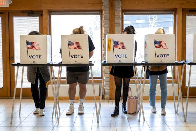 Four people stand in a row at a table, their faces and torsos obscured by white boxes that have American flags on it and say 'Vote.'