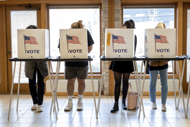 Four people stand in a row at a table, their faces and torsos obscured by white boxes that have American flags on it and say 'Vote.'