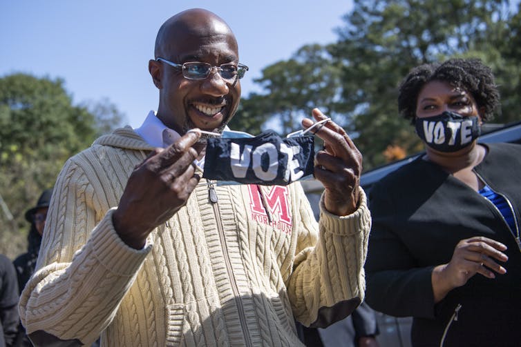 A middle aged Black man with glasses holds a black mask that says 'vote' in white. He stands next to a middle aged Black woman with a mask over her mouth that also says vote.