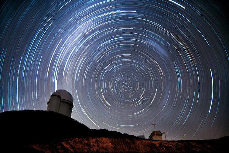 丘の中腹にあるドーム型望遠鏡のシルエットの背後にある夜空に星が円を描く様子を示す長時間露光の写真。