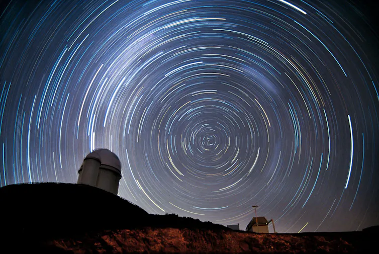A long-exposure photo showing stars tracing out circles in the night sky behind the silhouette of a domed telescope on a hillside.