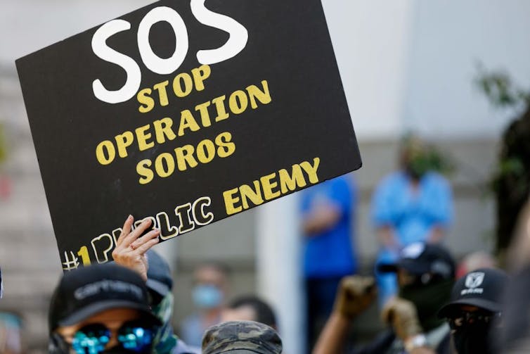 Someone holds up a poster that says 'SOS Stop Operation Soros #1 Public Enemy'