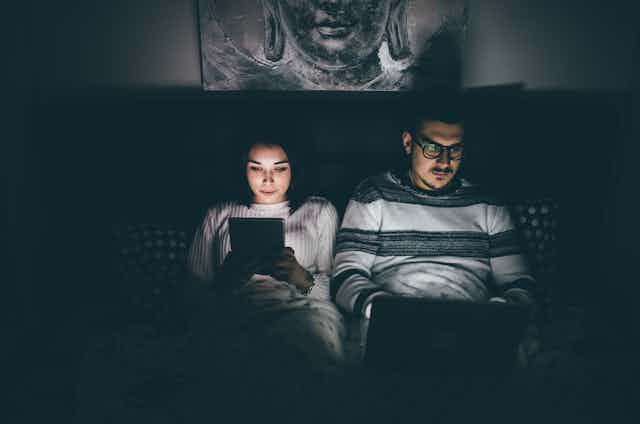A couple lief in bed in a dark room. Both people are on a digital device