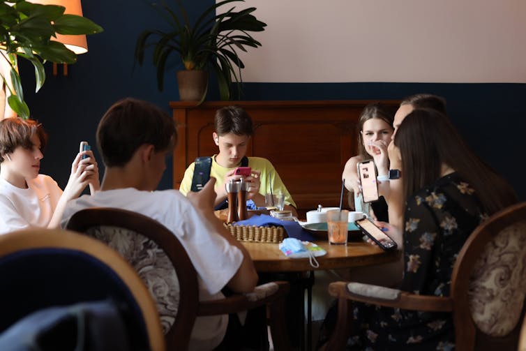 A group of young people sit at a round table, some occupied by their phones