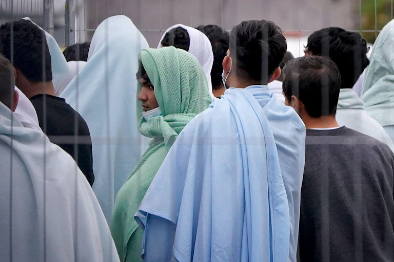 A group of men thought to be refugees, wrapped in sheets and blankets and standing behind a fence, turned away from the camera