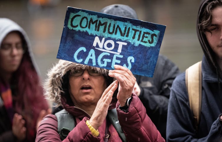 A demonstrator in a warm coat holds a handmade sign reading 'communities not cages'.