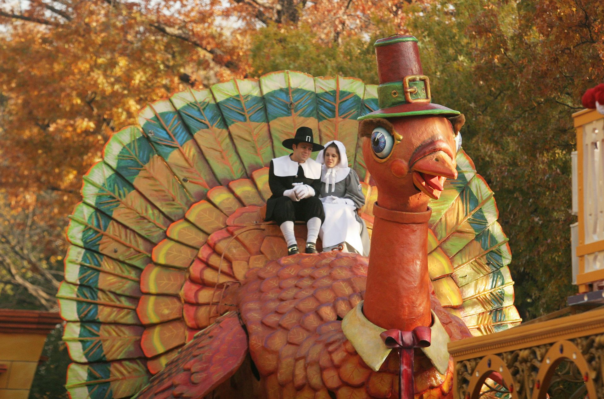 Two people dressed as Pilgrims sit atop a giant turkey float that is wearing a Pilgrim hat.