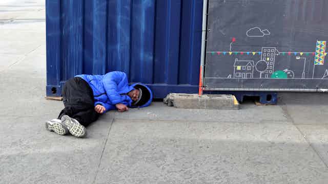 A man in a blue jacket and black trousers sleeps on a pavement in front of a blue container and a blackboard with a drawing of a house.