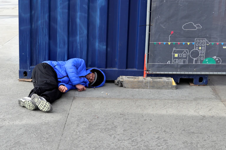 A man in a blue jacket and black trousers sleeps on a pavement in front of a blue container and a blackboard with a drawing of a house.