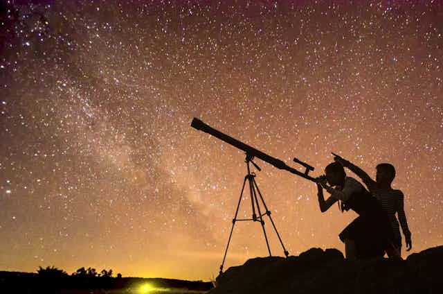 Silhouette of two children looking through telescope at Milky Way