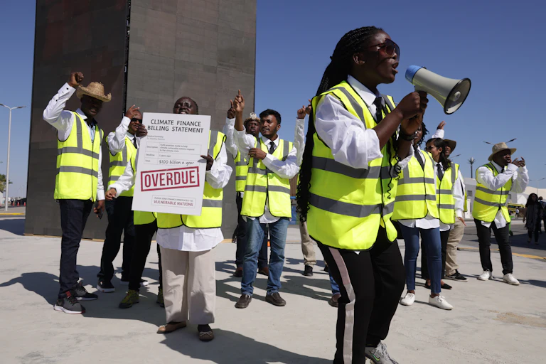 Person with loudspeaker leading others with placards and raised hands in a protest.