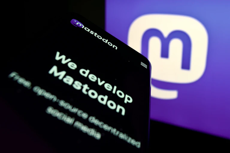 a screen showing the mastodon logo — a blue m in a white speech bubble - there's a phone screen with the text WE DEVELOP MASTODON showing