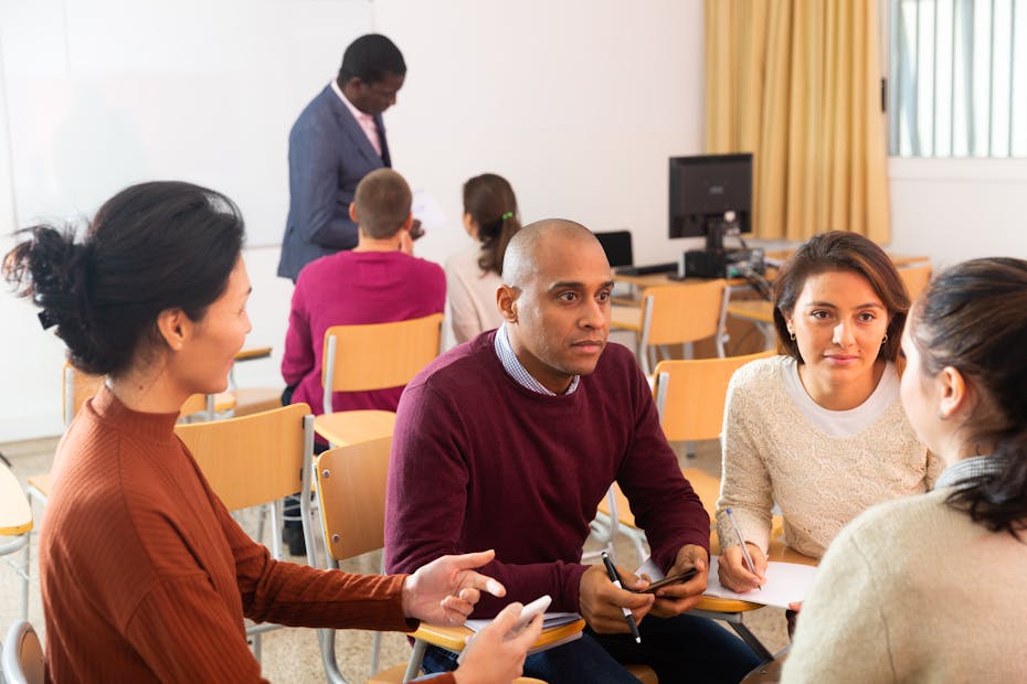 Three women and a man sit in a circle talking in a classroom.