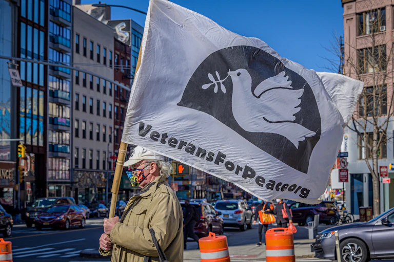 A white older man with a beige jacket holds a white flag that says Veterans for Peace dot org