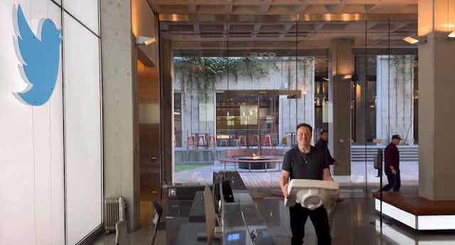 Elon Musk stands in a building lobby, holding a sink, near the Twitter logo