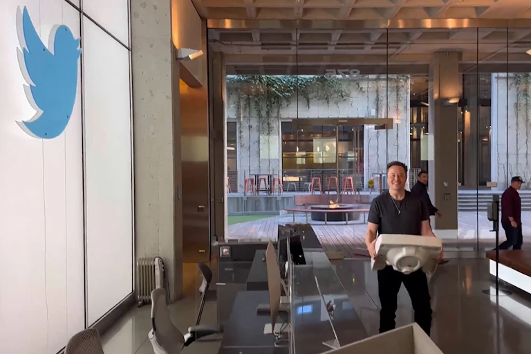 Elon Musk stands in a building lobby, holding a sink, near the Twitter logo