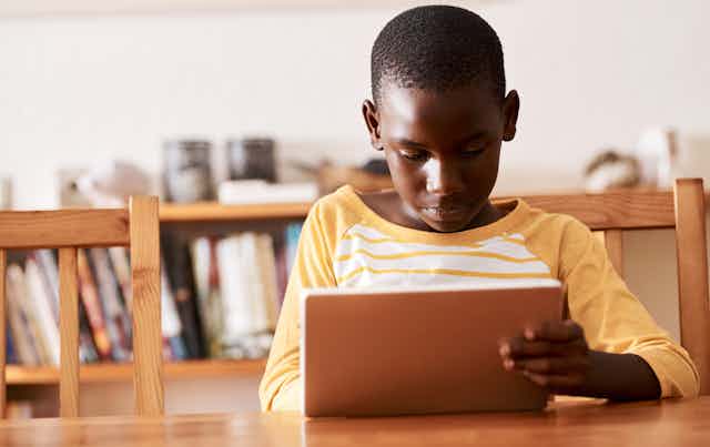 A Black boy wearing a yellow and white striped shirt holds a tablet while seated at a wooden table inside his home.