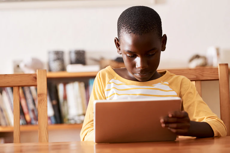 A Black boy wearing a yellow and white striped shirt holds a tablet while seated at a wooden table inside his home.