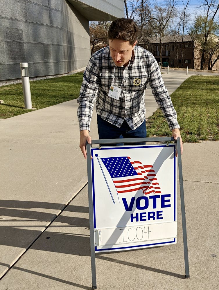 A man in a plaid shirt sets up a sign that has an American flag on it and the words 'VOTE HERE.'