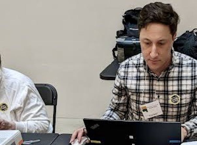 A dark-haried man in a plaid shirt working at a computer next to a printer.