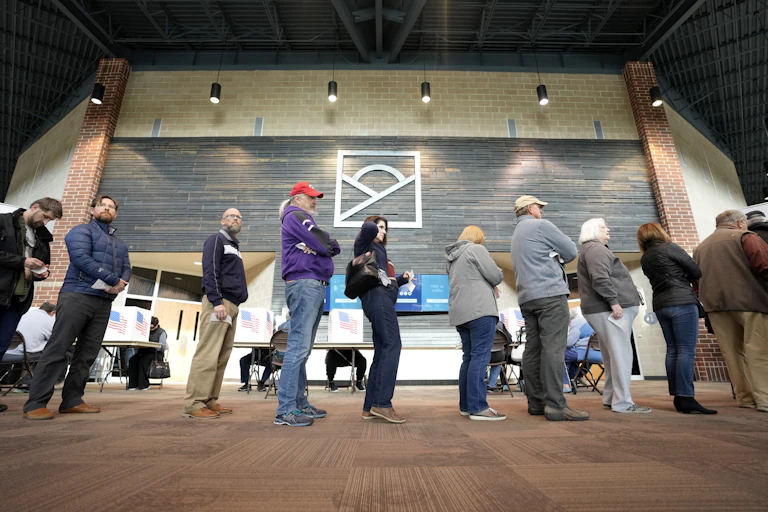Almost a dozen people stand in line to get their ballots in a large, public space.