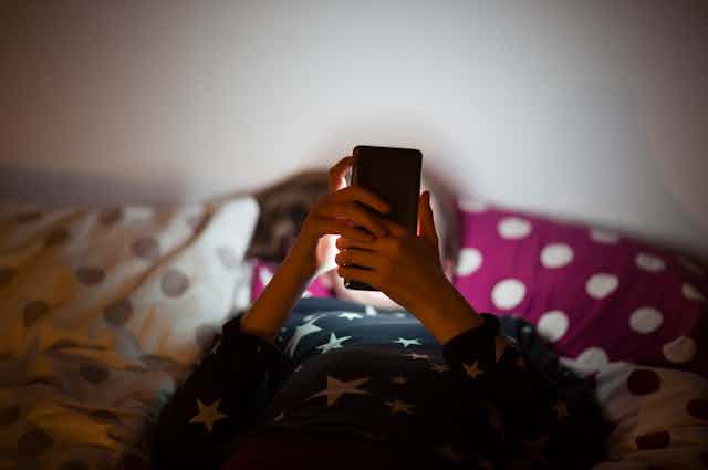 Kid in bed at night, face hidden by smartphone.
