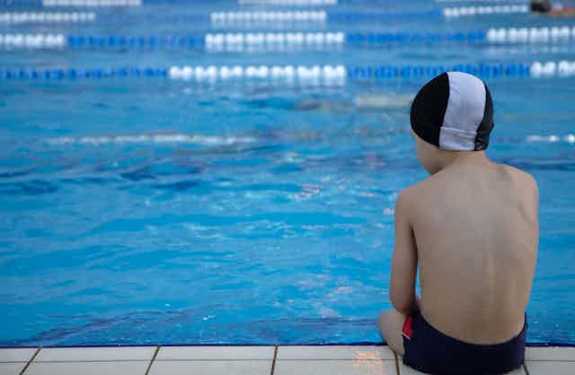 a child wearing swim trunks and a swim cap sits at the edge of a swimming pool