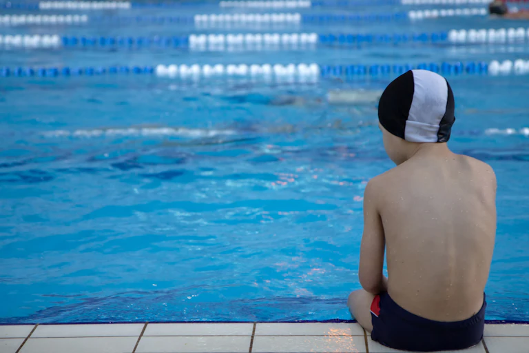 a child wearing swim trunks and a swim cap sits at the edge of a swimming pool